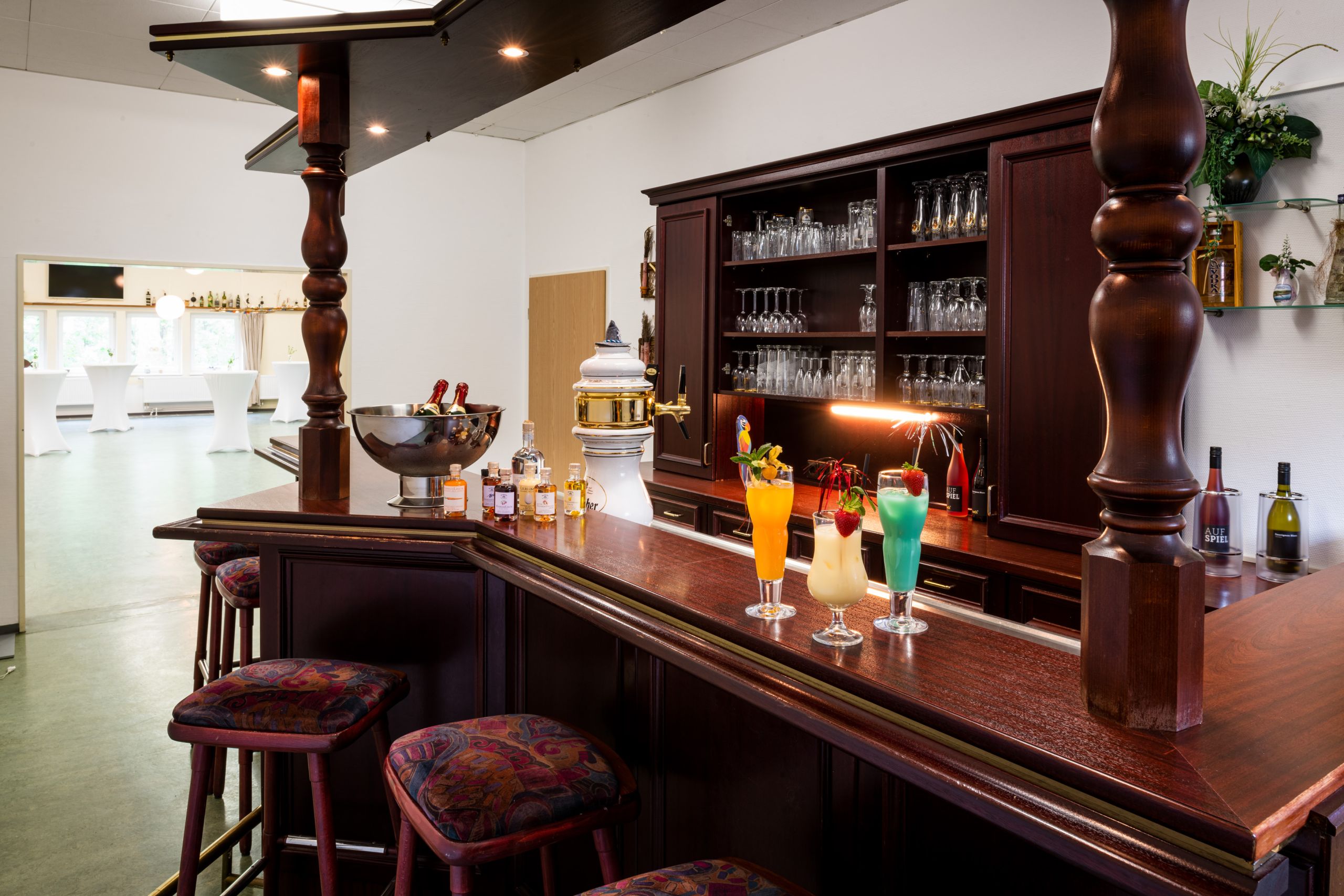 Auf einer Bar aus dunklem Holz stehen bunte Cocktails und Liköre, im Hintergrund stehen Gläser im Schrank.