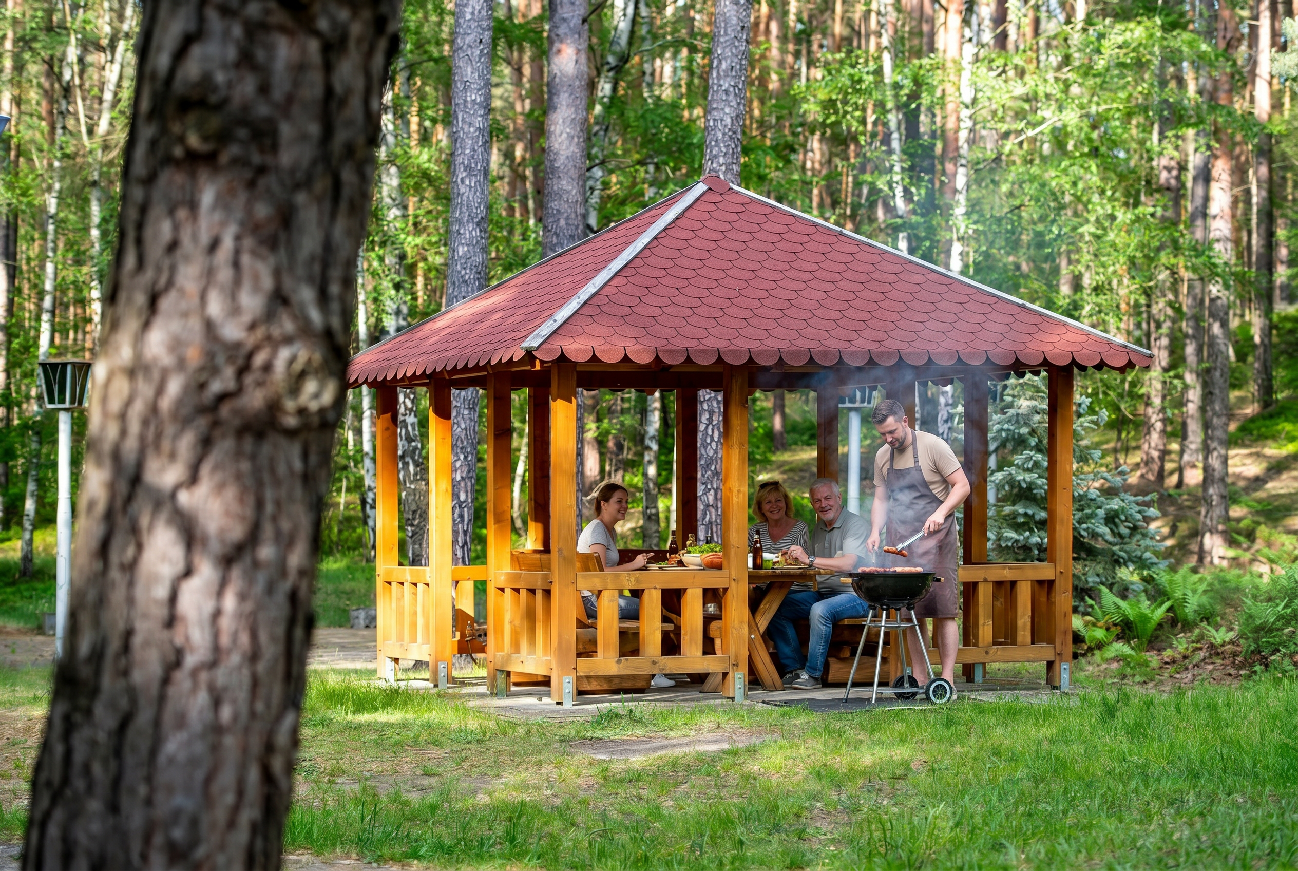 Eine Personengruppe sitzt in einer Holzhütte, davor steht eine Person am grill